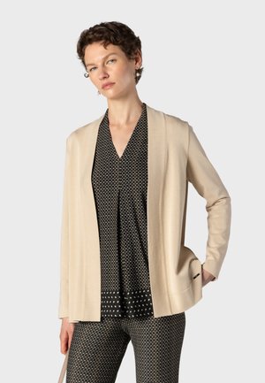Femme aux cheveux courts et bouclés porte un cardigan beige ouvert sur un top noir à motifs et un pantalon assorti sur un fond gris uni.