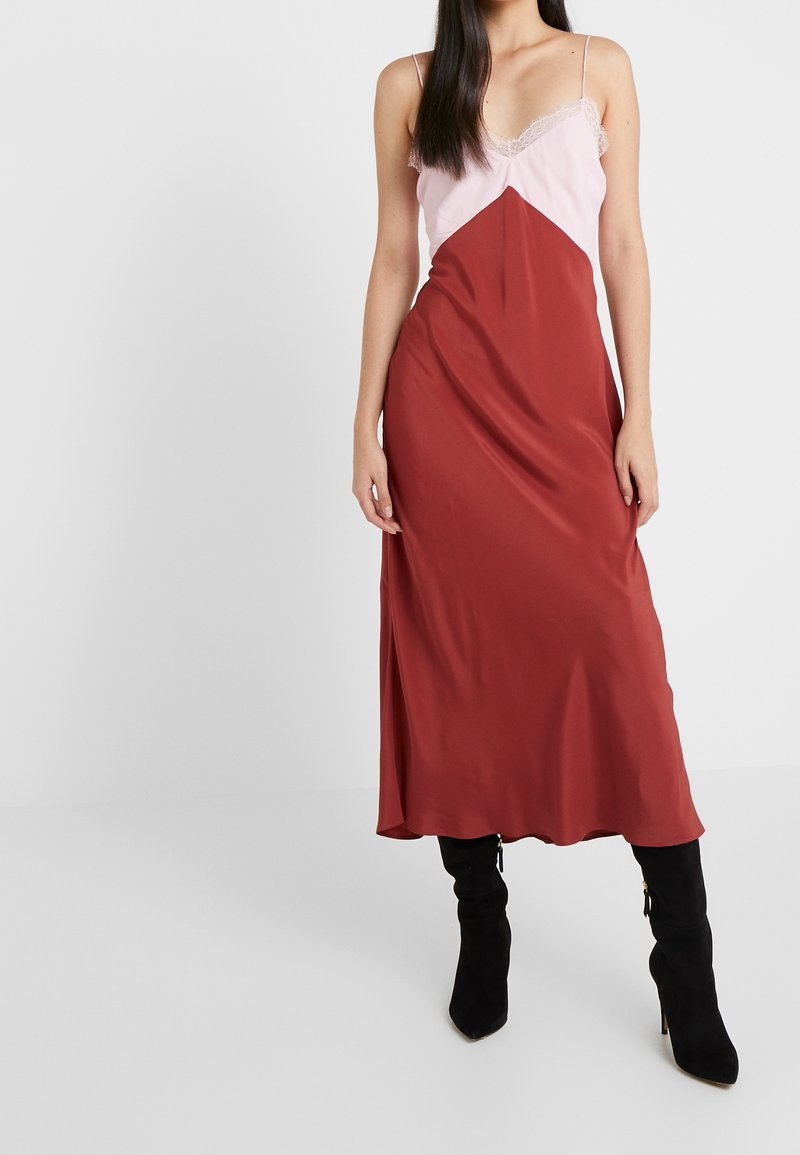 Femme portant une robe nuisette sans manches rose et rouge associée à des bottes à talons noires, debout devant un fond blanc uni.
