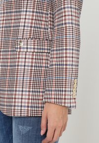 Blazer en motif écossais rose, bleu et marron ; doté d'une poche poitrine unique et de quatre boutons crème sur le revers.