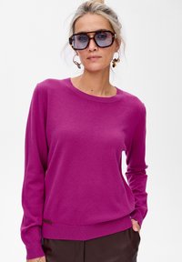 Kobieta z blond włosami związanymi w kok, nosząca oversizedowe okulary w kolorze żółwia, kolczyki w kształcie okręgów w tym samym kolorze, magentowy sweter i ciemne spodnie.