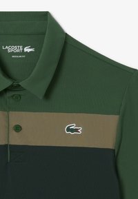Grön polo skjorta med mörka och olivgröna horisontella ränder, Lacoste krokodil-logotyp på bröstet, knäppt krage och "Lacoste Sport"-etikett inuti kragen.