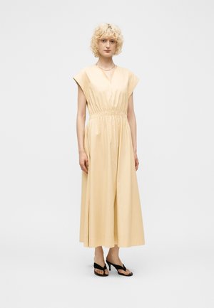 Femme aux cheveux blonds bouclés portant une robe midi beige à manches courtes et des sandales noires ouvertes à talons, debout devant un fond uni.