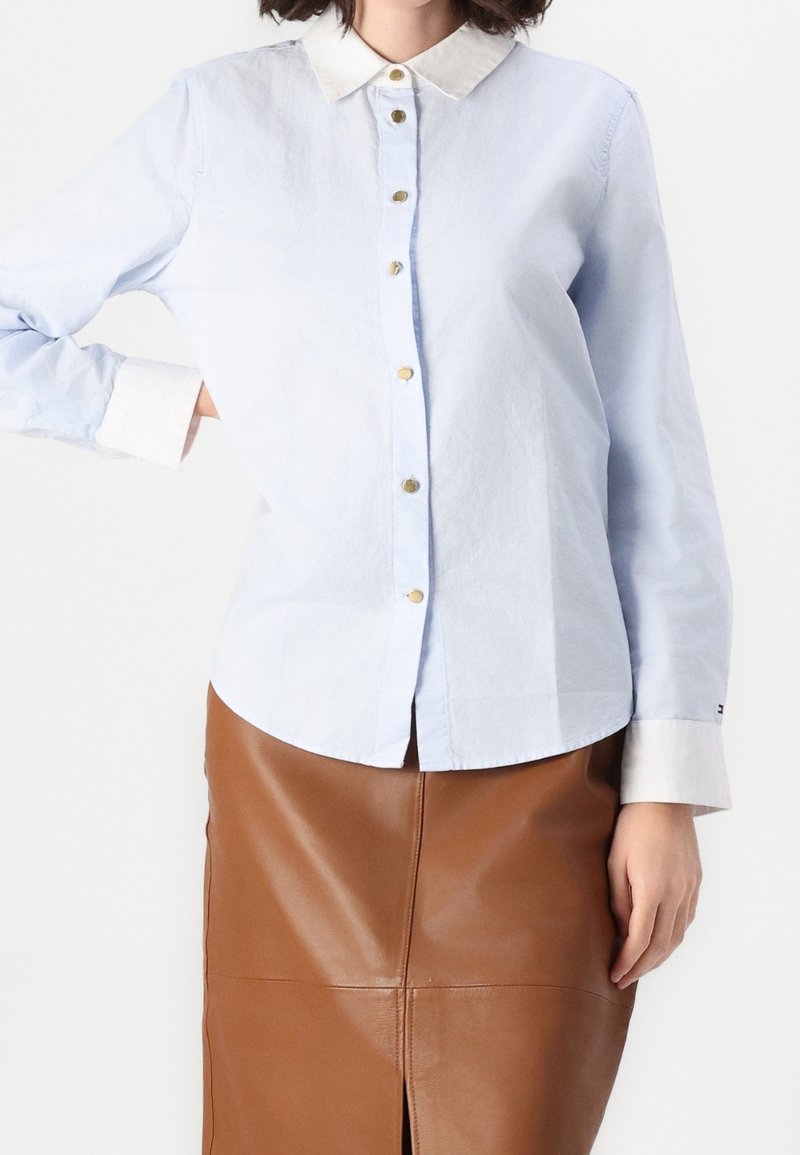 Una donna che indossa una camicia azzurro chiaro con bottoni, colletto e polsini bianchi, abbinata a una gonna in pelle marrone fino al ginocchio.