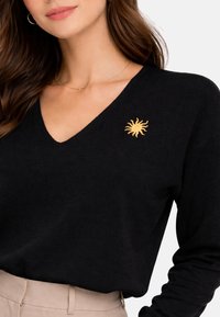 Femme portant un pull noir à col en V avec une petite broderie de soleil jaune sur la poitrine et un pantalon beige taille haute, vue de l'épaule à la taille.