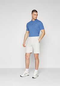 Camisa polo azul com colarinho e três botões, combinada com calções brancos. Usada com ténis brancos com detalhes cinzentos.