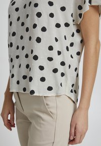 Blusa leggera e semi-trasparente con orlo arrotondato, caratterizzata da pois neri su uno sfondo crema. Il tessuto ha una finitura testurizzata.