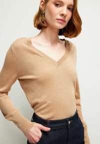 Beige V-Ausschnitt-Pullover aus weichem Stoff, mit langen Ärmeln und gerippten Bündchen, kombiniert mit dunkelblauen hochgeschnittenen Jeans.