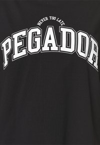 Svart t-shirt i bomull med en djärv vit "PEGADOR" grafisk design och texten "NEVER TOO LATE" i en böjd layout ovanför.