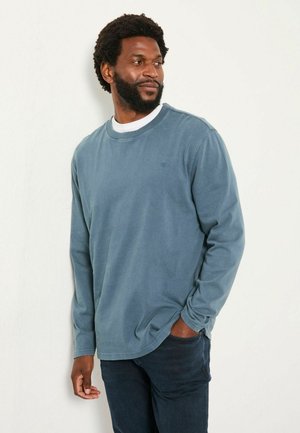 Mann mit Bart, der ein blaues Langarmhemd über einem weißen T-Shirt und dunklen Jeans trägt, steht mit einer Hand in der Tasche und schaut zur Seite gegen eine weiße Wand.