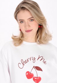 Felpa bianca con una stampa rossa di due ciliegie e il testo "Cherry Me". Il modello indossa orecchini a forma di ciliegia rossi, con una texture morbida.