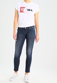 Vit t-shirt med kort ärm och "DIESEL"-logotyp, kombinerad med mörkblå slim fit jeans och silverfärgade sneakers, som visar upp en avslappnad stil.