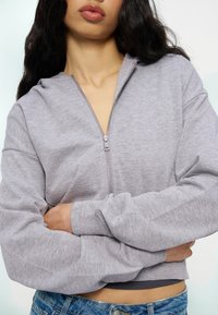 Femme croisant les bras, portant un sweat à capuche gris clair zippé et un jean bleu, montrant le ventre contre un fond uni.