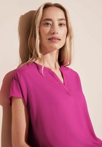 Blusa fucsia con scollo a V, maniche corte e una texture liscia. Il tessuto appare leggero e fluido su uno sfondo neutro.