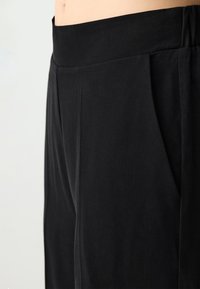 Zwarte broek met een tailored ontwerp, een gladde textuur, ingestikte plooien aan de voorkant en zijzakken. De tailleband lijkt breed en elastisch.