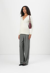 Cremfarbener V-Ausschnitt-Langarmtop, kombiniert mit lockeren grauen Hosen und schwarzen spitzen Ballerinas. Das Model hält eine bordeauxfarbene Handtasche und steht vor einem weißen Hintergrund.
