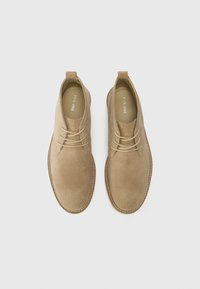 Botines de ante beige con punta redonda, diseño de cordones, acentos de costura y lengüetas en el talón. Suela plana, con un branding mínimo en el interior.