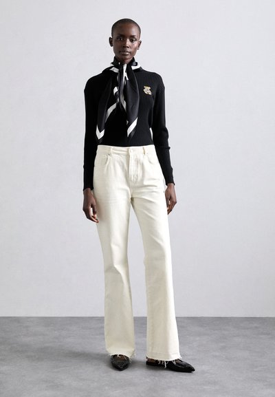 Maglione nero a coste con spilla, pantaloni crema a gambe larghe e sciarpa nera e bianca con motivo a fantasia. La modella indossa scarpe nere a punta.