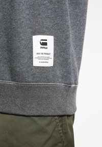 Sudadera gris que presenta una etiqueta tejida con el logo "G STAR RAW", textura lisa, puños acanalados y dobladillo recto. Combinada con pantalones verde oliva.