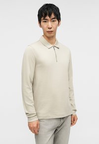 Μπεζ πόλο μπλούζα με μακριά μανίκια και φερμουάρ στο γιακά, κατασκευασμένη από λείο ύφασμα, συνδυασμένη με ανοιχτό γκρι παντελόνια.