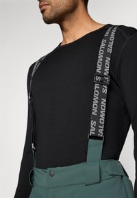 Macacão impermeável verde com suspensórios ajustáveis pretos, apresentando a marca "SALOMON" e uma textura suave. Parte superior preta de manga longa visível.