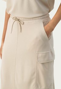 Jupe en coton beige avec une taille élastique et un cordon de serrage, des poches latérales, et une texture lisse avec un design simple.