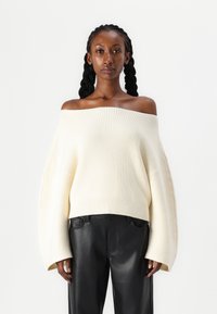 Pull en tricot ivoire dénudé d'une épaule avec larges manches et texture côtelée, accompagné d'un pantalon en cuir noir.