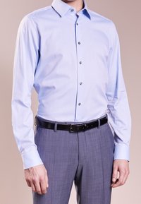 Světle modrá košile s dlouhým rukávem a límečkem, která má šest kovových knoflíků a hladký povrch, spárovaná se šedými vzorovanými kalhotami a černým opaskem.