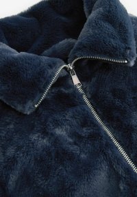 Jachetă din blană artificială de culoare bleu marin, cu un finisaj moale și texturat. Dispune de un fermoar argintiu, guler zimțat și un design pufos, confortabil.