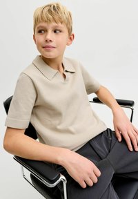 Polo beige à manches courtes en tissu texturé avec un col classique, associé à un pantalon foncé, assis sur une chaise noire.