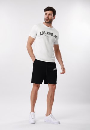 Homme portant un T-shirt blanc avec l'inscription « Los Angeles », un short noir avec le texte « Los Angeles » et des baskets blanches, debout devant un fond blanc uni.