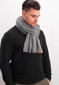Gray knitted scarf featuring a textured ribbed pattern, accompanied by a small leather tag. Worn around the neck over a black sweater.