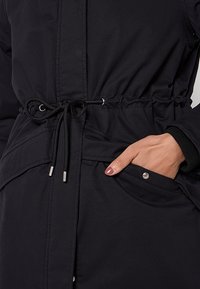 Zwarte parka met elastische taille, trekkoord, zijzakken en zilverkleurige hardware. Glad textuur en lichtgewicht stof.
