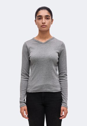 Jeune personne aux cheveux foncés portant un haut gris à manches longues avec col en V et un pantalon noir, debout devant un fond clair uni.