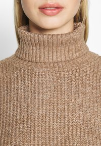 Tan ribbad polokrage-tröja med hög krage och texturerad stickad mönster, tillverkad av mjukt material med en mysig känsla.