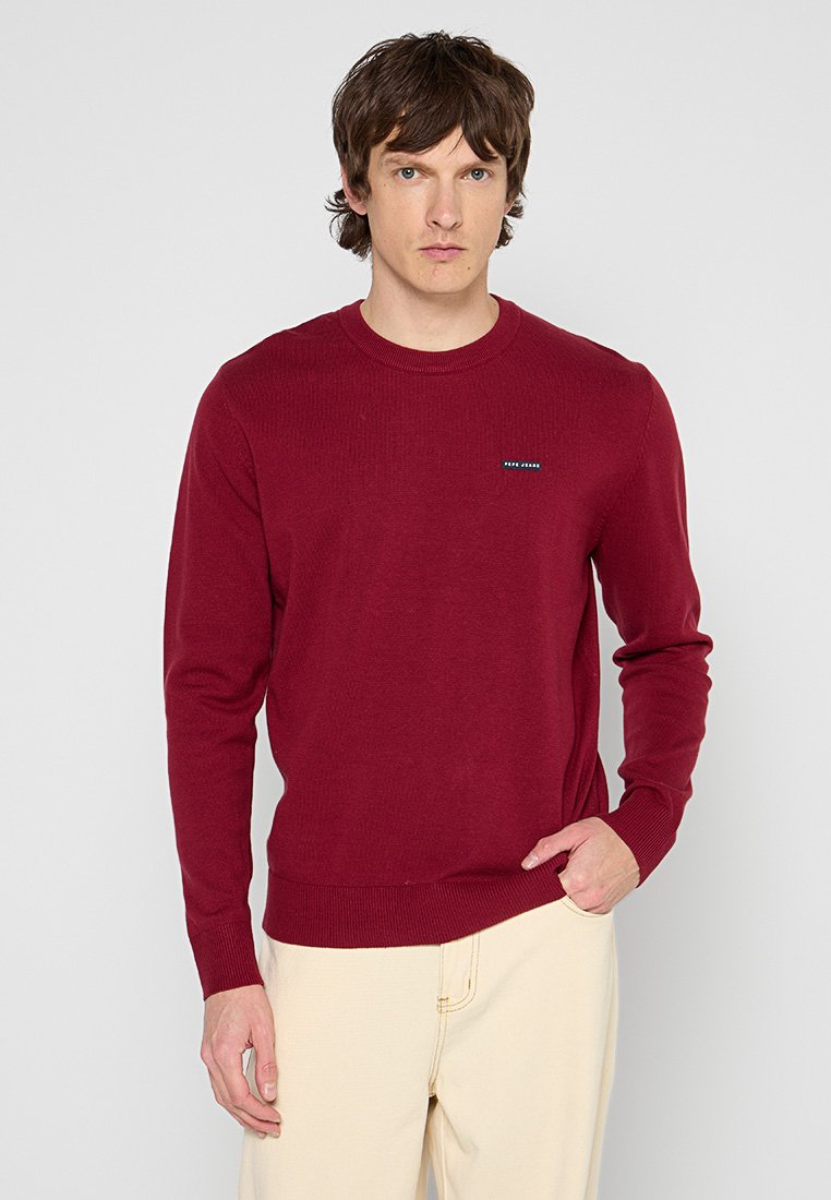 Pull en maille bordeaux, avec un col rond, des poignets côtelés et un petit logo en accent sur la poitrine.