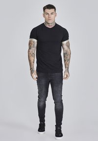 Svart åtsittande t-shirt med kort ärm och en kontrasterande gyllene detalj vid ärmsluten, parat med mörka tight jeans och svarta sneakers.