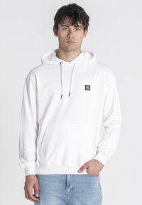 Sudadera blanca con capucha, con un bolsillo canguro frontal, cordones ajustables y un parche de logo negro en el pecho. Textura de tela suave.