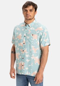 Camicia a maniche corte con colletto di colore azzurro chiaro, decorata con un motivo floreale astratto in rosa, dotata di taschino sul petto e bottoni neri. Realizzata in tessuto leggero.