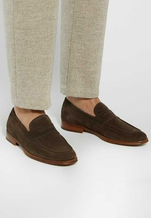 Brown suede loafers with a low heel, featuring stitching details and a smooth leather insole. Worn with light beige tailored pants.
