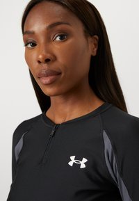Black athletic top with a partial zipper, featuring a gray accent on the shoulder and a white logo on the chest. Smooth texture.