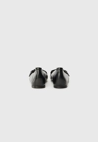 Chaussures en cuir noir avec une forme arrondie, présentant une texture lisse et un design minimaliste. Deux sangles réglables avec boucles sur les côtés.