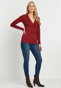 Maglietta bordeaux a maniche lunghe con scollo a V e design avvolgente, abbinata a jeans skinny blu e stivaletti marroni.