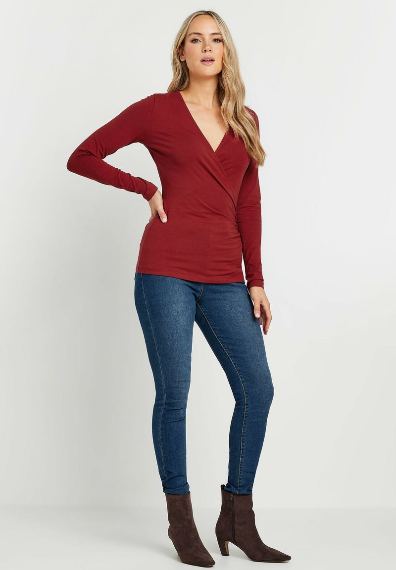 Maglietta bordeaux a maniche lunghe con scollo a V e design avvolgente, abbinata a jeans skinny blu e stivaletti marroni.