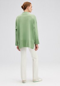 Pulover oversized verde deschis, cu mâneci lungi, tiv ondulat, slituri laterale, asortat cu pantaloni albi și adidași albi. Textură netedă.