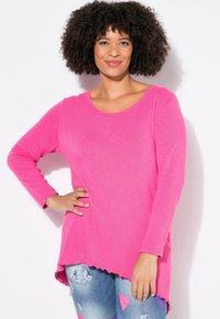 Helles pinkes Langarmoberteil mit runder Ausschnitt und einem längeren, welligen Saum. Kombiniert mit zerrissenen blauen Jeans mit Herzmustern.
