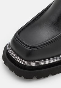 Chaussure en cuir noir avec semelle épaisse et détails à clous argentés autour du bord près de la zone des orteils.