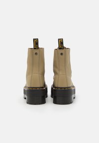 Stivali ankle in pelle beige con suole nere spesse, cuciture gialle e linguette nere con testo giallo, mostrati da dietro su uno sfondo bianco.