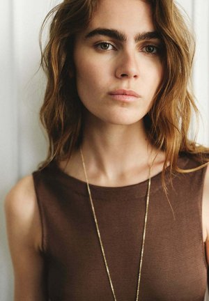 Joven mujer con cabello ondulado castaño claro, que lleva una camiseta marrón sin mangas y un collar delgado de oro, mirando ligeramente más allá de la cámara.