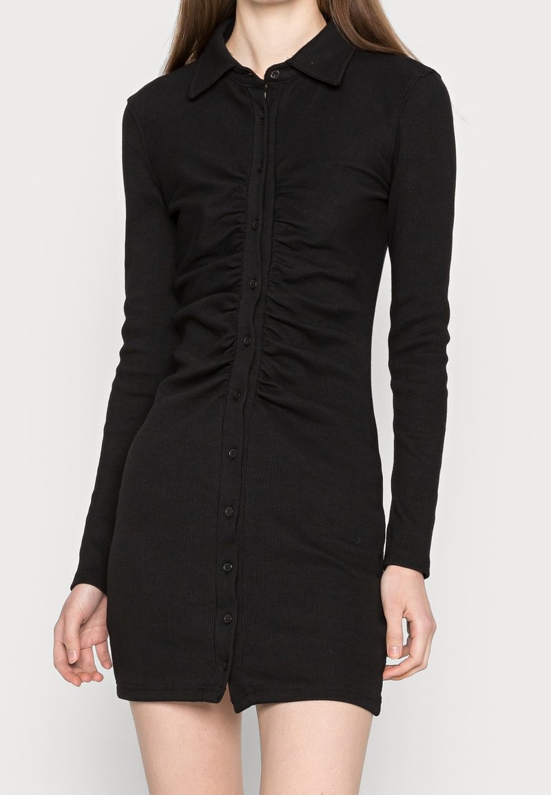 Svart ränsad långärmad klänning med knäppning framtill, som har rynkade detaljer längs bålen och en figurnära silhuett.