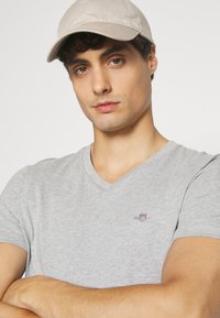 Siva v-neck majica s malim logotipom na prsima, uparena s bež kapom. Tkanina izgleda glatko, a kroj je uski.
